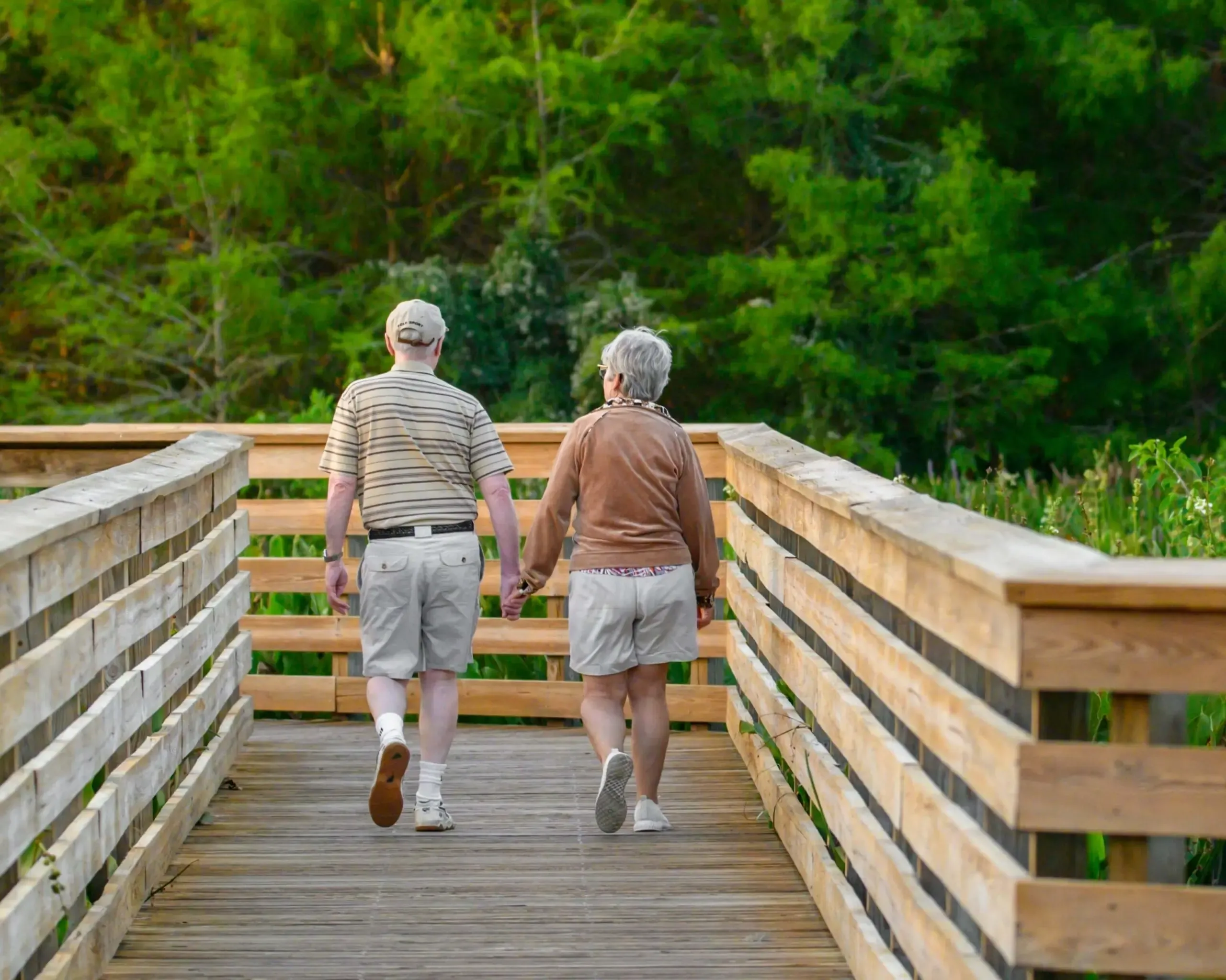 An older couple walking hand in hand along a wooden boardwalk through a green Florida wetland.